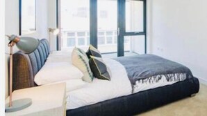 A bedroom with a bed, a bedside table with a lamp, and large windows. - Impeccable 1-bed Apartment in London (London)