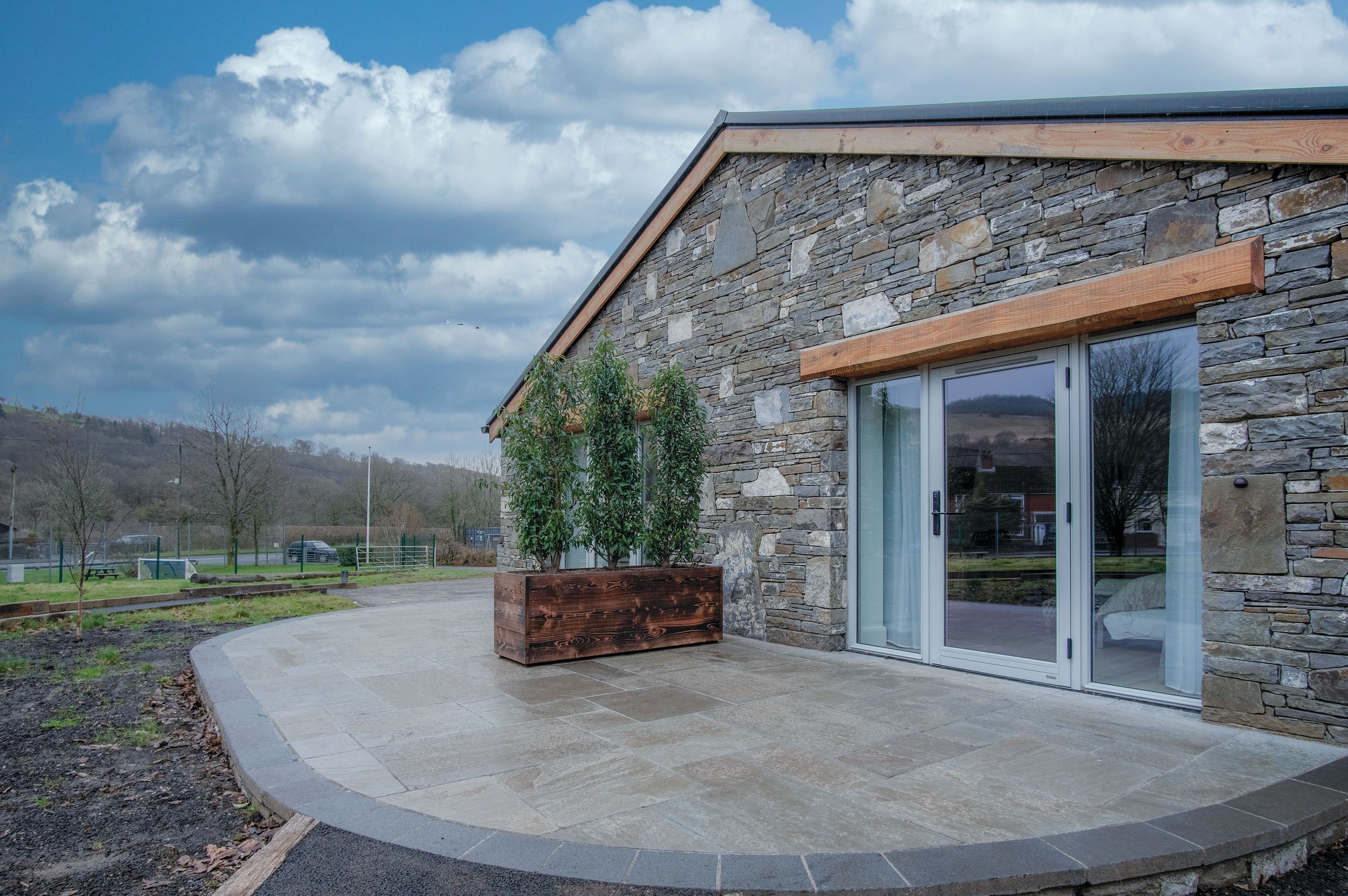 A modern building with stone exterior, large sliding glass doors, and a wooden planter with greenery.