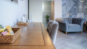 A wooden dining table with a woven basket containing snacks and a water bottle. A grey armchair and a small side table with a lamp are visible in the background. - The Sleeping Giant - Pen Y Cae Inn (Swansea)