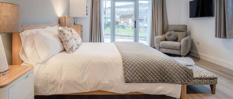 Un dormitorio moderno con una cama grande, una silla, un televisor y un ventanal corredero al exterior.