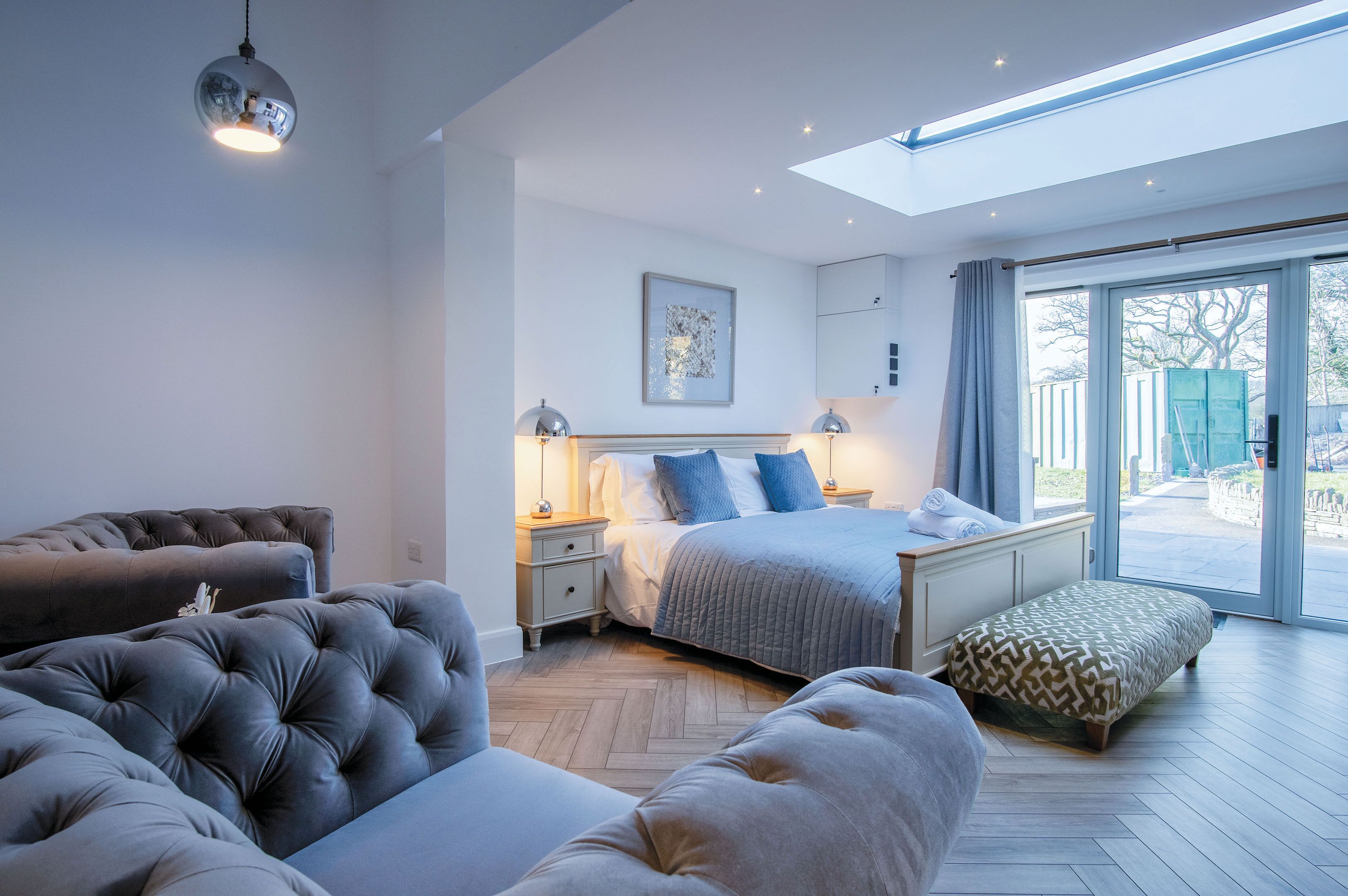 Ein modernes Schlafzimmer mit einem großen Bett, einer Couch und einer Bank. Über dem Bett befindet sich ein Oberlicht und durch die Glastüren hat man einen Ausblick nach außen.