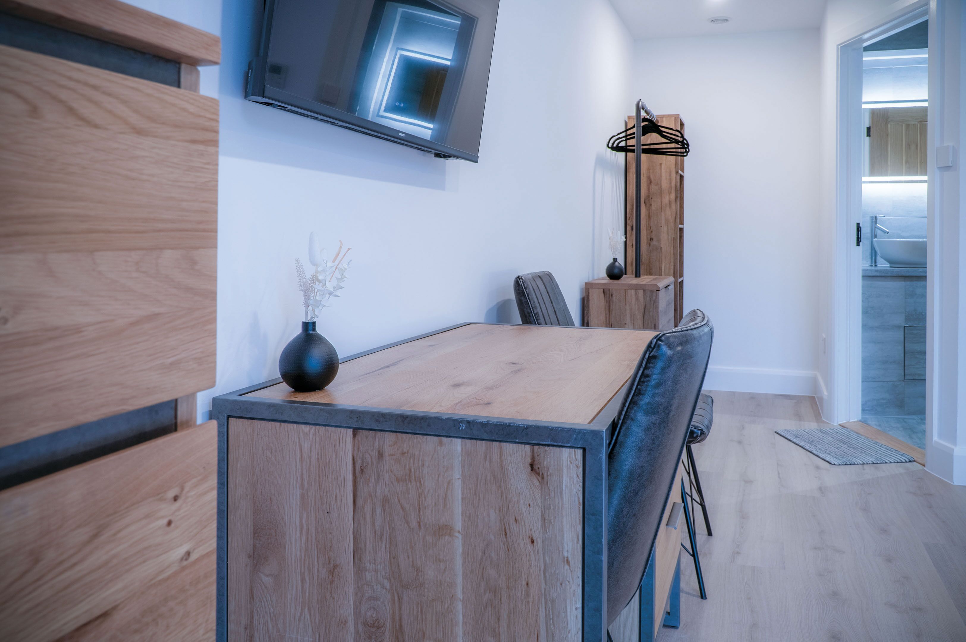 A modern kitchen with a wooden table, chairs, a TV mounted on the wall, and a view into the bathroom.
