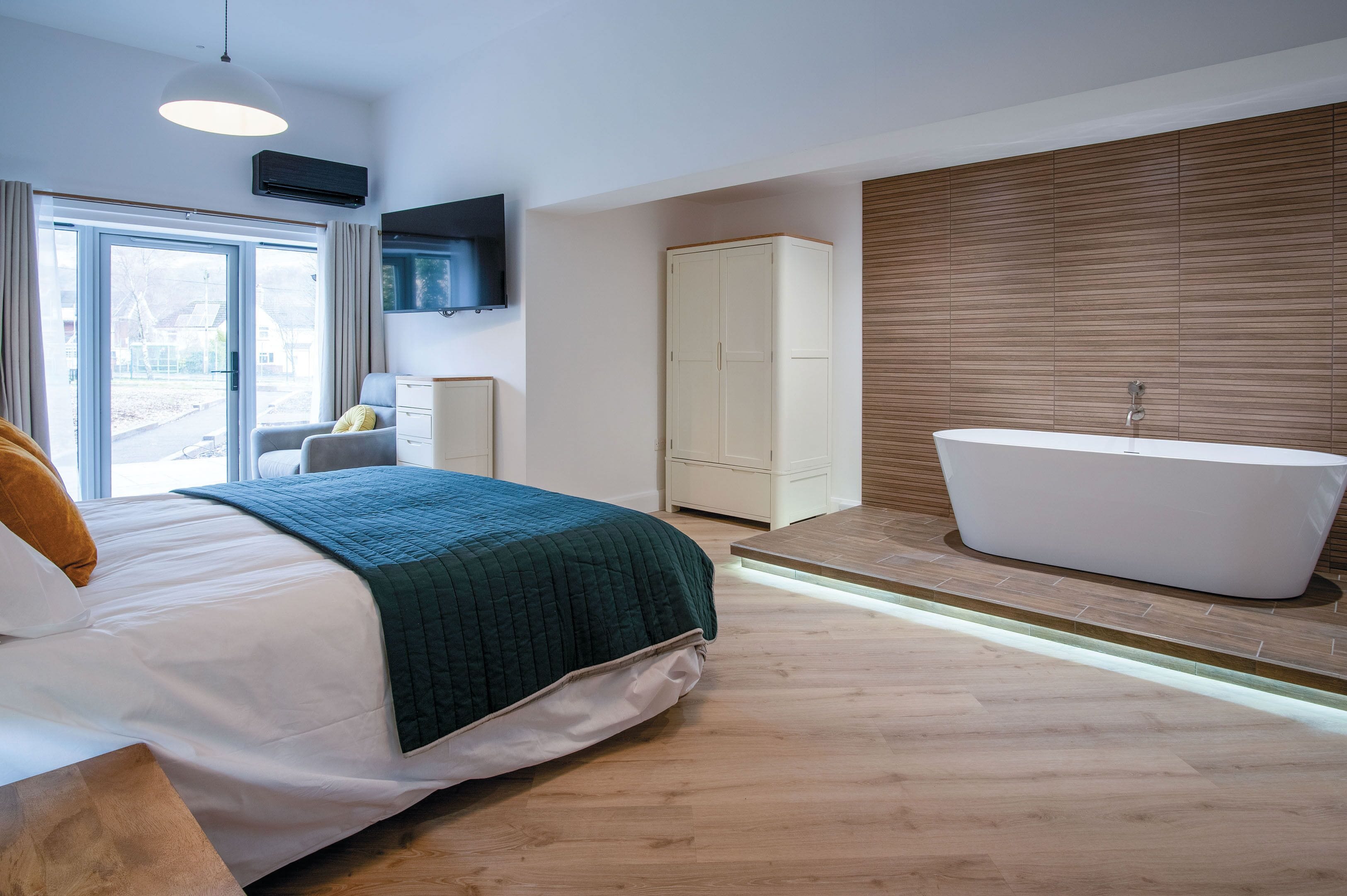 A modern bedroom with a large bed, a built-in bathtub, and a view through a sliding door.