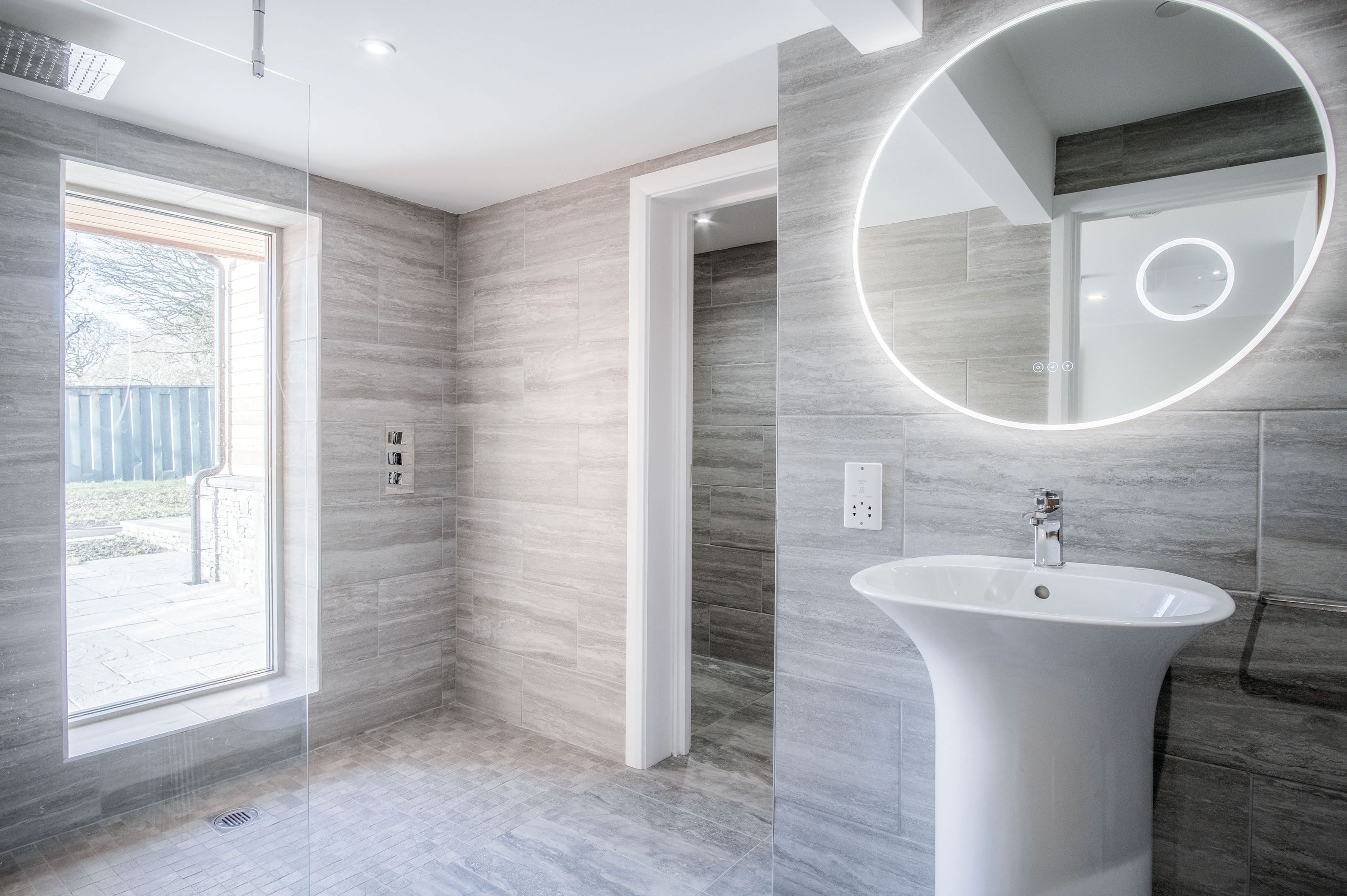 A modern bathroom with a large window, a round mirror, and a pedestal sink.