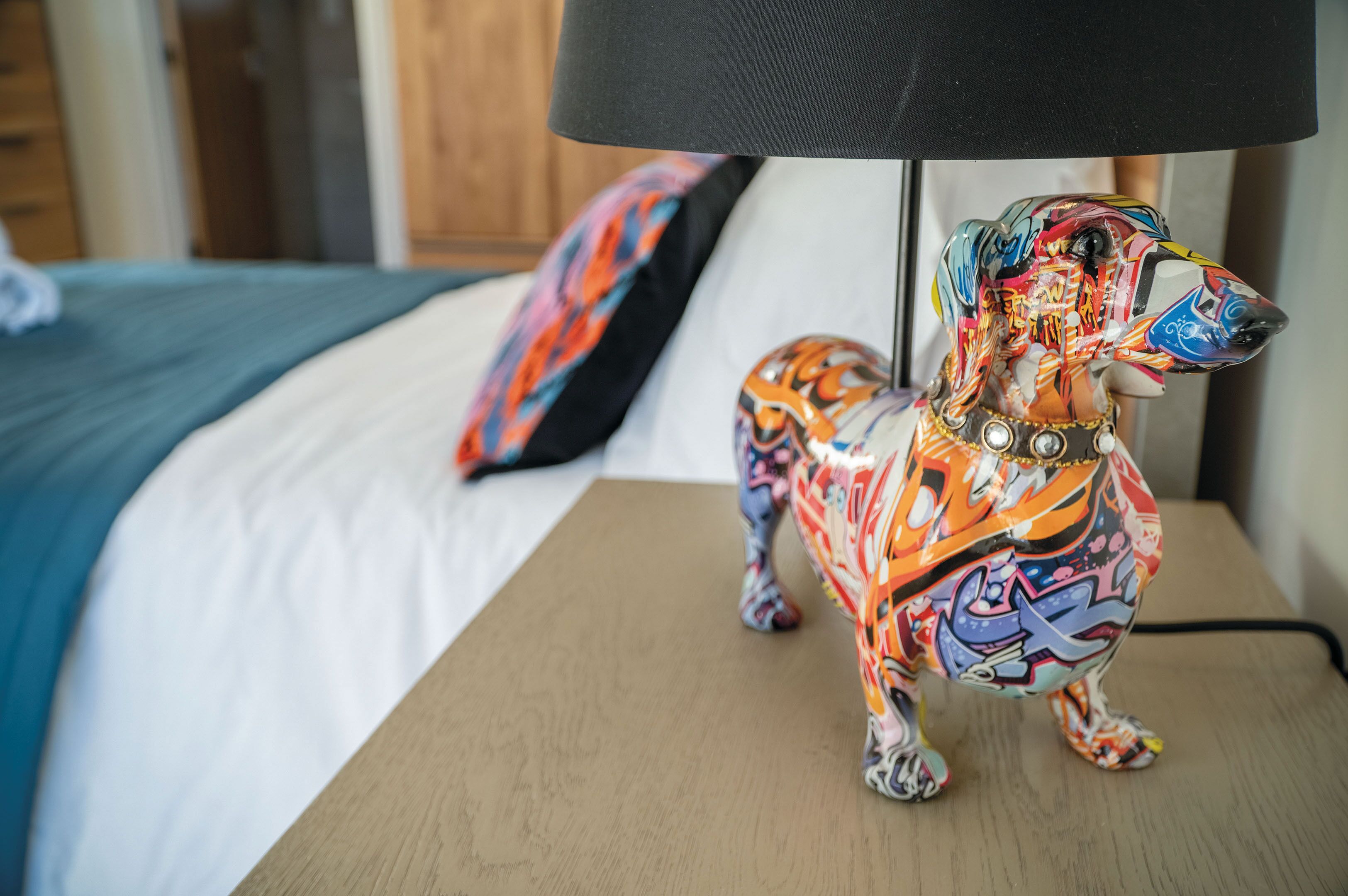 Eine farbenfrohe Hunde-Figur auf einem Nachttisch mit einer schwarzen Lampe, ein Bett mit einer blau-weißen Tagesdecke und ein buntes Kissen in einem Hotelzimmer.