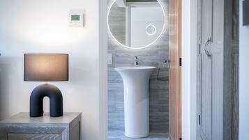 A modern bathroom with a round mirror, a white sink, and a lamp on a wooden nightstand.