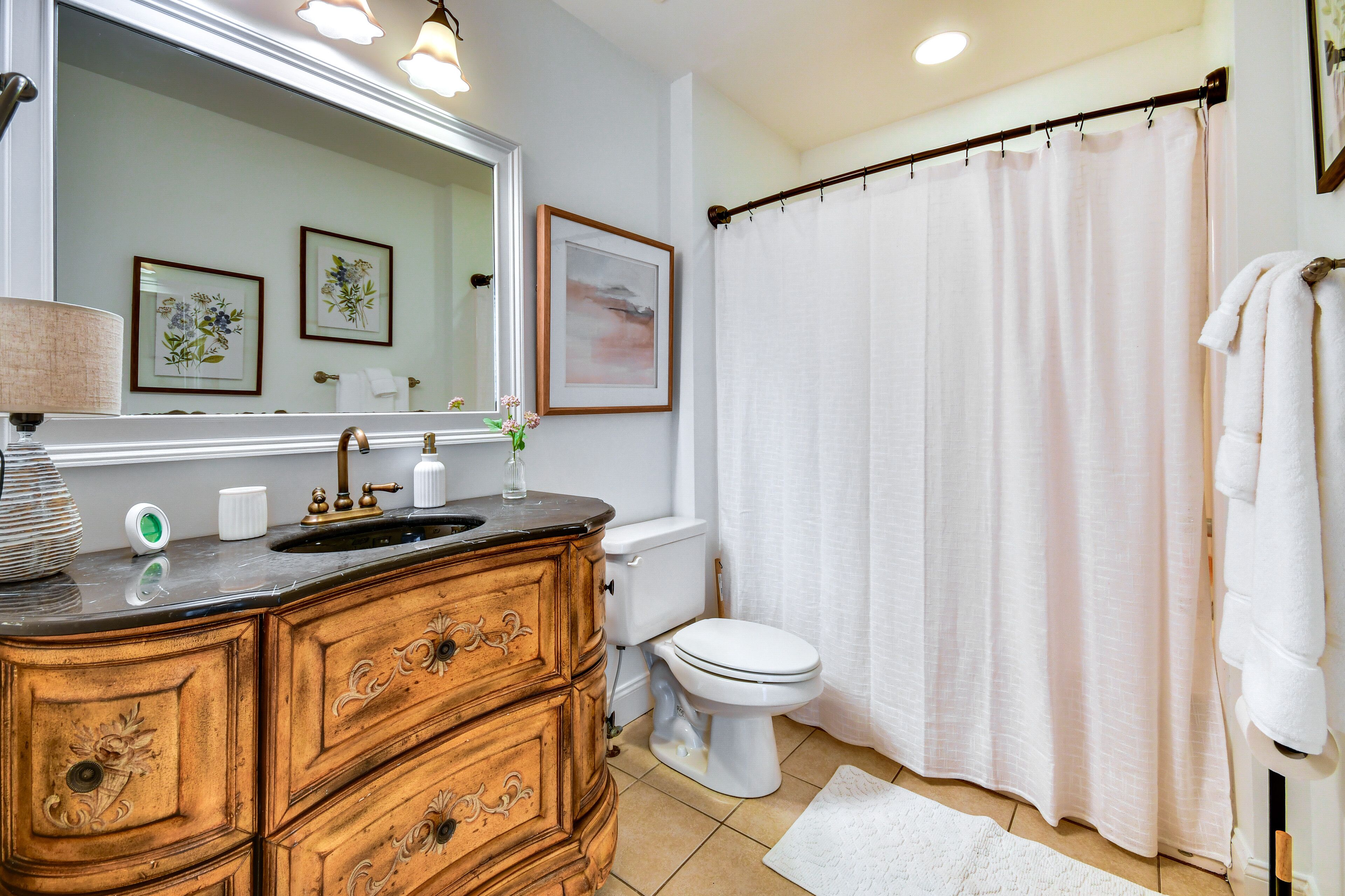 A bathroom with a wooden vanity, a white toilet, a shower curtain, and framed artwork on the wall.