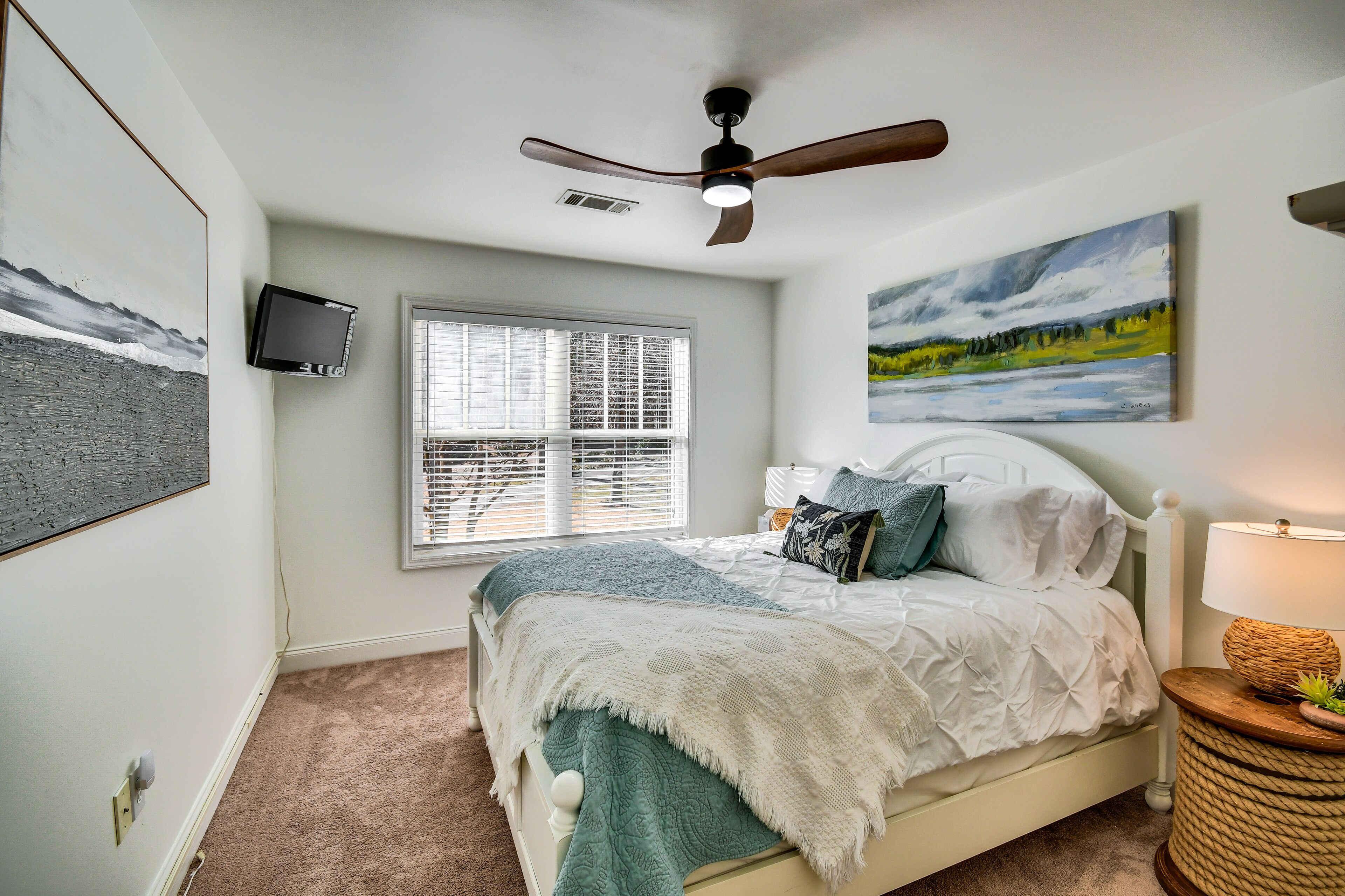 A bedroom with a bed, a ceiling fan, a window, a painting, and a lamp.