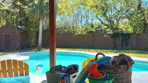 Un espace aménagé au bord de la piscine, avec une piscine, une chaise en bois, une table sur laquelle se trouvent divers objets, et une clôture en arrière-plan.