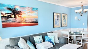 A living room with a gray sofa, a dining table with chairs, and a large painting of a sunset over the ocean. - Amazing Oceanfront Condo, Corner Unit, Natural Lights, 4th Floor (Myrtle Beach)