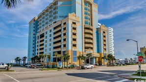 A multi-story hotel with a blue and beige facade, located at a street corner with palm trees and a stop sign. - Amazing Oceanfront Condo, Corner Unit, Natural Lights, 4th Floor (Myrtle Beach)