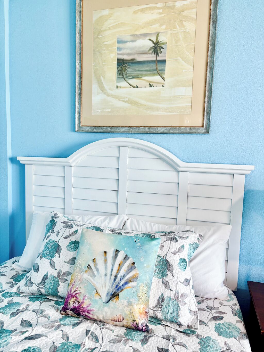 A bedroom with a white headboard, a bed with floral bedding, and a framed picture of a beach scene on the wall.