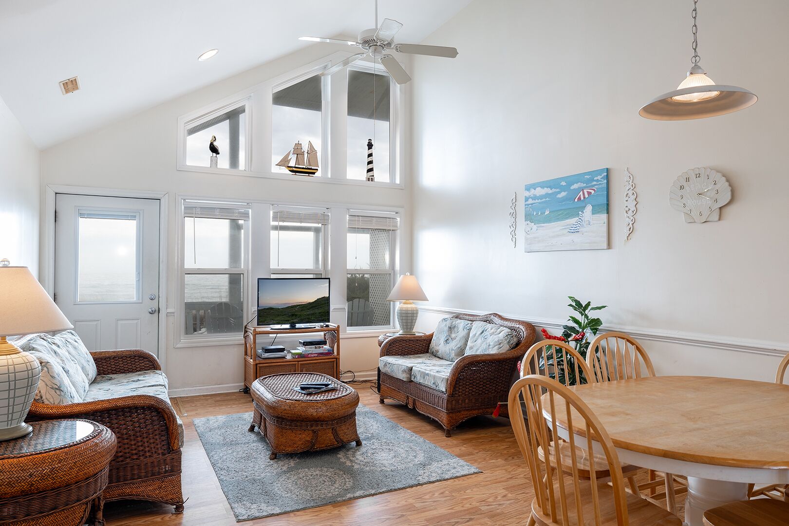 A spacious living room with a high ceiling, large windows, wicker furniture, a dining table, and a television.