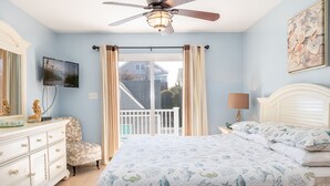 A bedroom with a bed, a dresser, a chair, a ceiling fan, a TV, and a balcony with curtains.