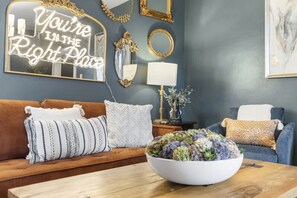 Un salon avec un canapé marron, des coussins blancs et à motifs, une table basse en bois agrémentée d’un centre de table floral, un miroir mural avec un cadre doré et un panneau décoratif.