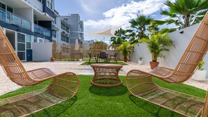 Un área de estar moderna al aire libre con sillas de mimbre y una mesa redonda, rodeada de césped artificial y plantas en macetas.