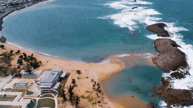 Vista aérea de una zona costera con playa arenosa, afloramientos rocosos y una zona residencial con edificios y vegetación.