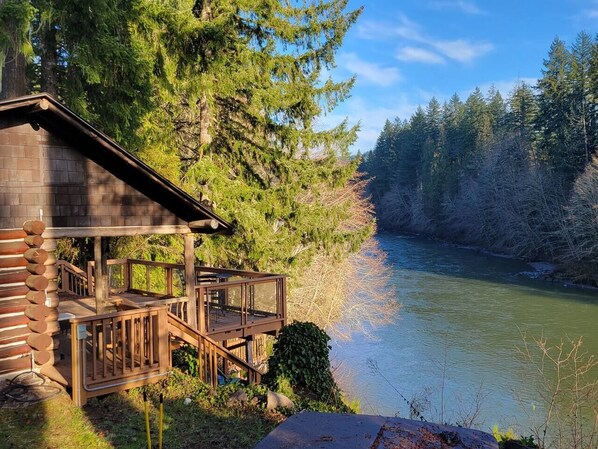 Une cabane dotée d’une terrasse donnant sur une rivière, entourée d’une forêt dense.