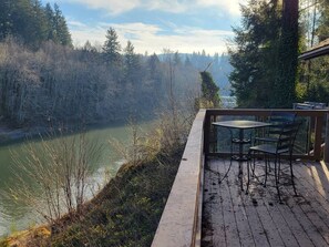 Une terrasse en bois avec une table et des chaises, donnant sur une rivière et entourée d’arbres.