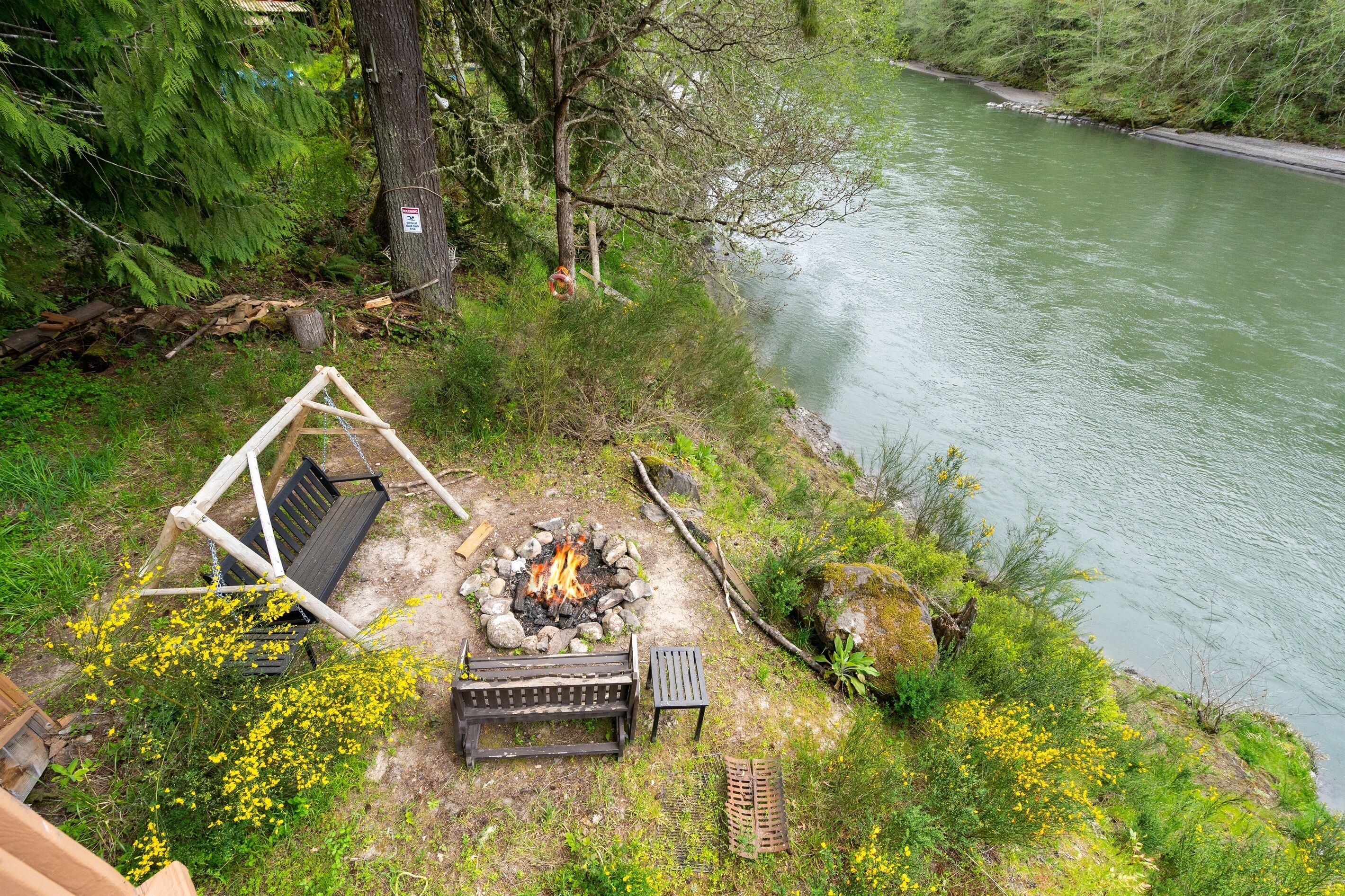 Un área de estar al aire libre junto al río, con un brasero, bancos y una mecedora.