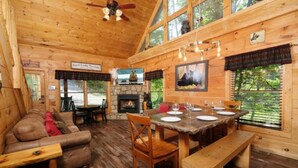 A cozy cabin interior with a stone fireplace, wooden dining table set for four, and a leather sofa. - Centrally Located between Pigeon Forge and Gatlinburg.  (Sevierville)
