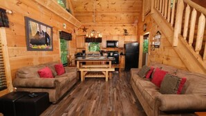 A cabin interior with two sofas, a wooden dining table, a kitchen area, and a staircase. - Centrally Located between Pigeon Forge and Gatlinburg.  (Sevierville)
