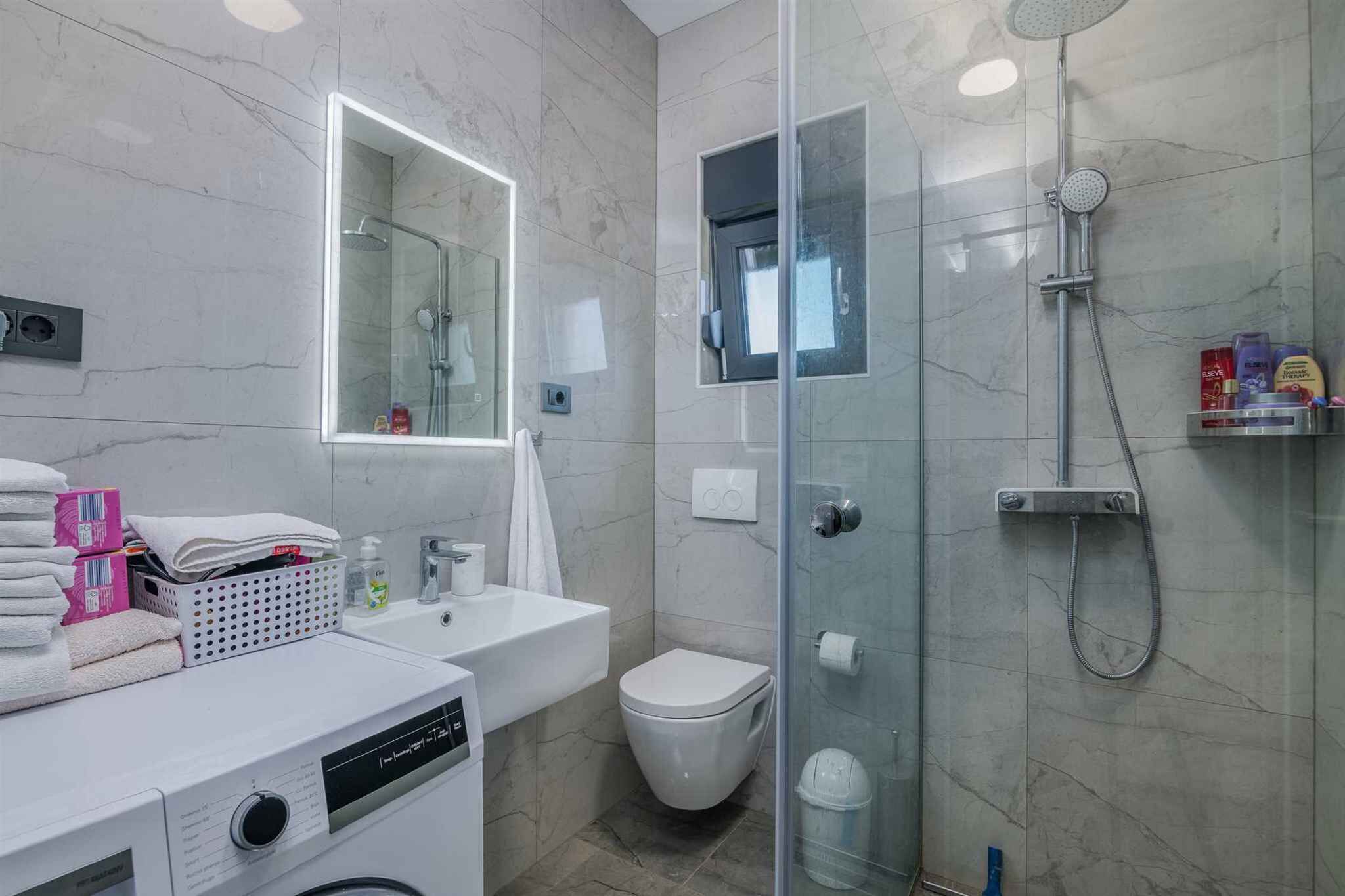 A modern bathroom with a glass-enclosed shower, a white sink, a toilet, and a laundry machine.