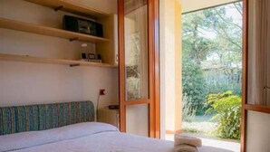 A bedroom with a bed, a window with a view of greenery, a shelf with items, and a ceiling light.