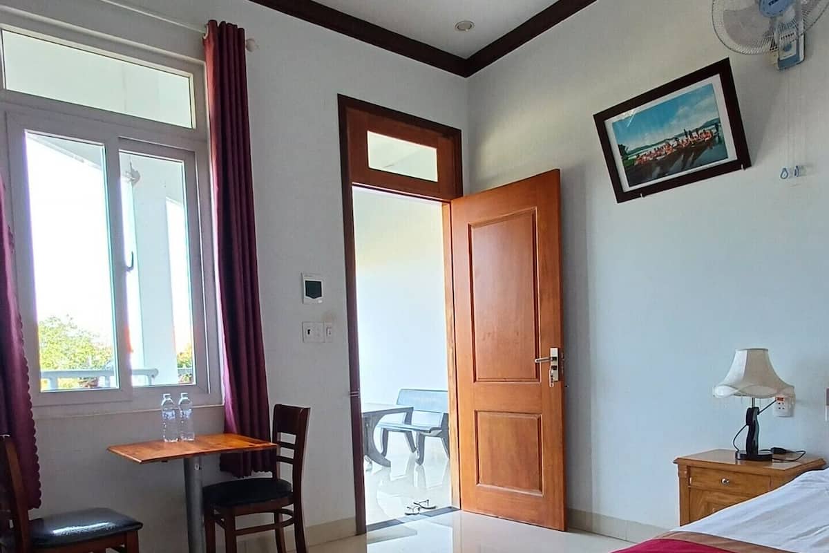 Quarto de hotel com cama, escrivaninha com cadeira, mesa com cadeiras, porta e janela com cortinas.