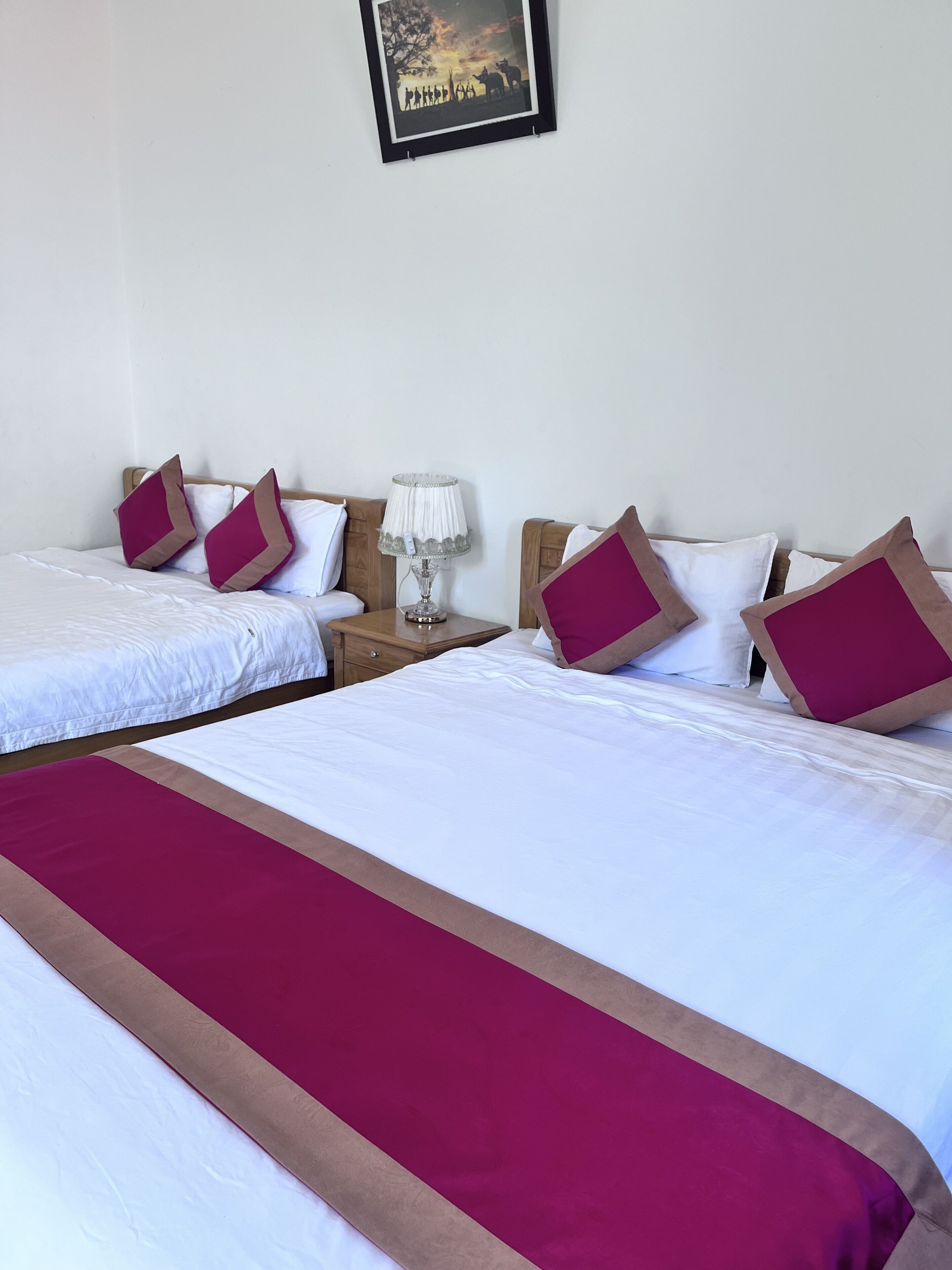 Habitación de hotel con dos camas, cada una con ropa de cama blanca y almohadas color burdeos. Entre las camas, un mesita de noche con una lámpara.