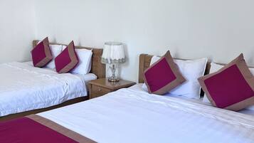 Habitación de hotel con dos camas, cada una con ropa de cama blanca y almohadas color burdeos. Entre las camas, un mesita de noche con una lámpara.