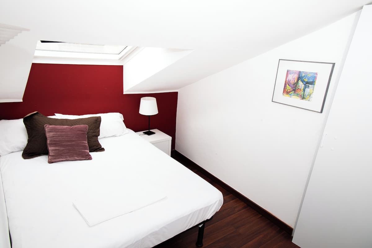 Cama bem arrumada com roupa de cama branca e marrom, parede de destaque vermelha, mesinha de cabeceira pequena com abajur e uma obra de arte emoldurada na parede.