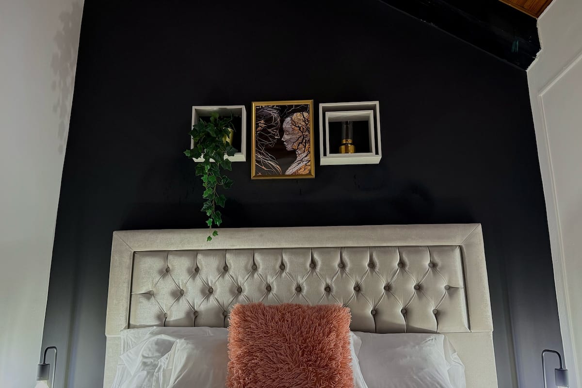 Quarto com cabeceira estofada, cama branca e almofada rosa fofa, além de três quadros na parede.