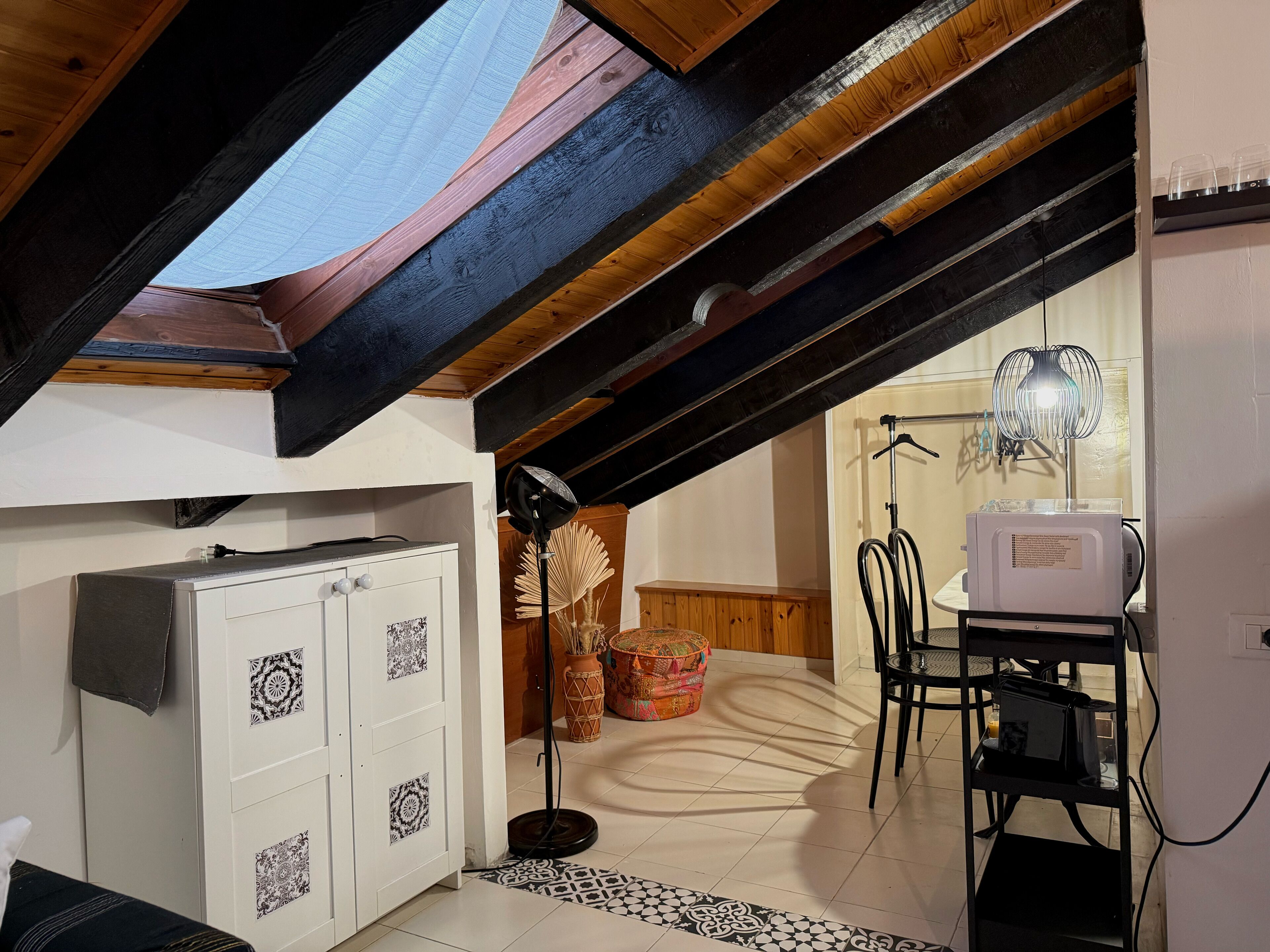 Une petite pièce mansardée, chaleureuse, avec un plafond en pente, une armoire blanche aux portes à motifs, une petite table ronde surmontée d’un coussin coloré, une chaise noire et un radiateur blanc.