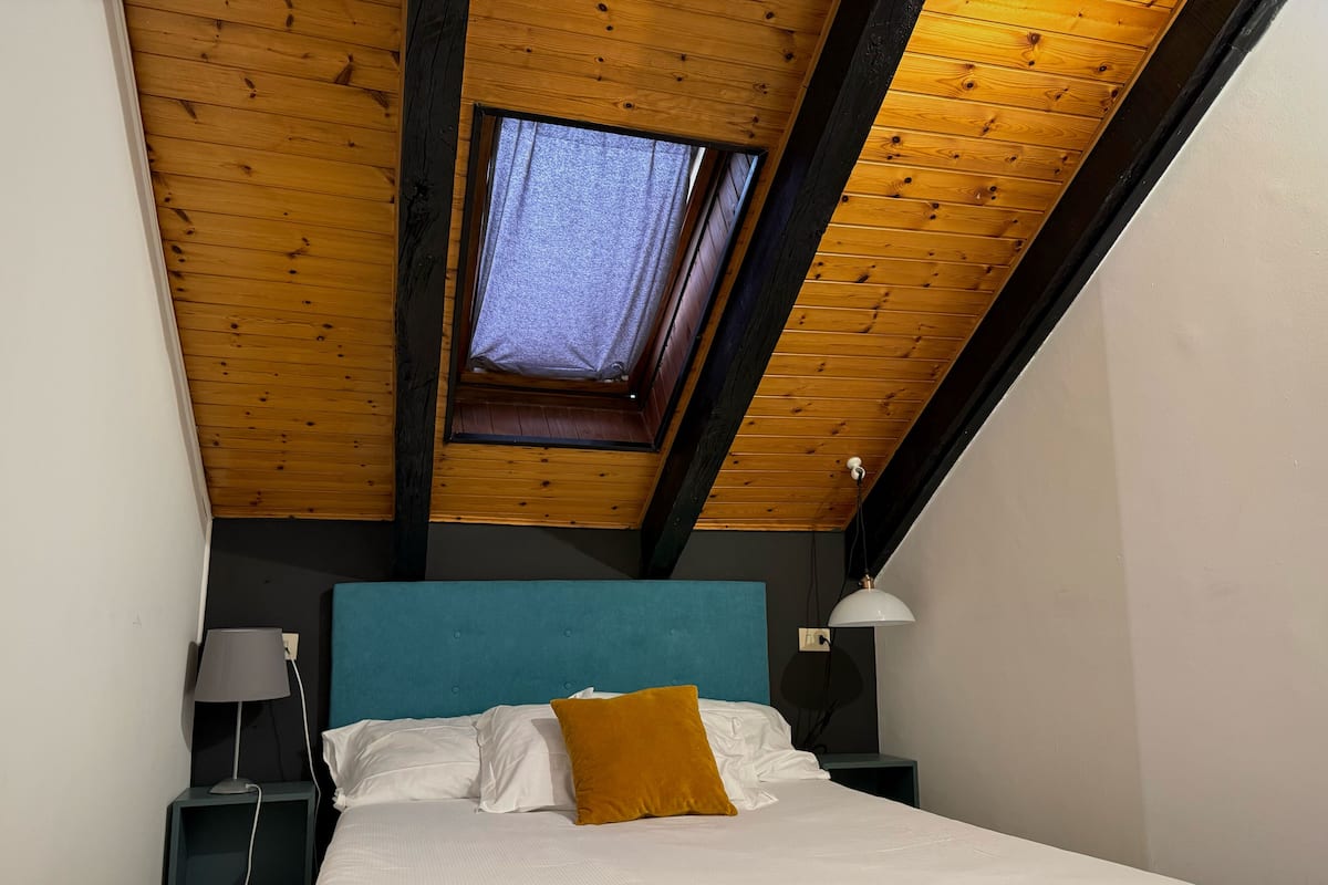 Quarto com teto inclinado, claraboia, cama com roupa de cama branca, cabeceira azul, travesseiro marrom e duas mesas de cabeceira com abajures.