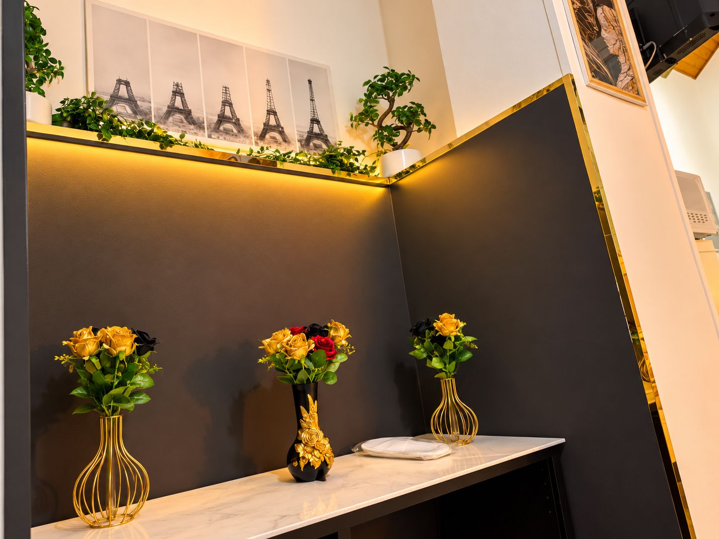 Ein modernes Interieur mit Marmorarbeitsplatte, drei Vasen mit Blumen, einem an der Wand befestigten Regal mit Pflanzen und einem gerahmten Bild, sowie einer dunklen Wand.