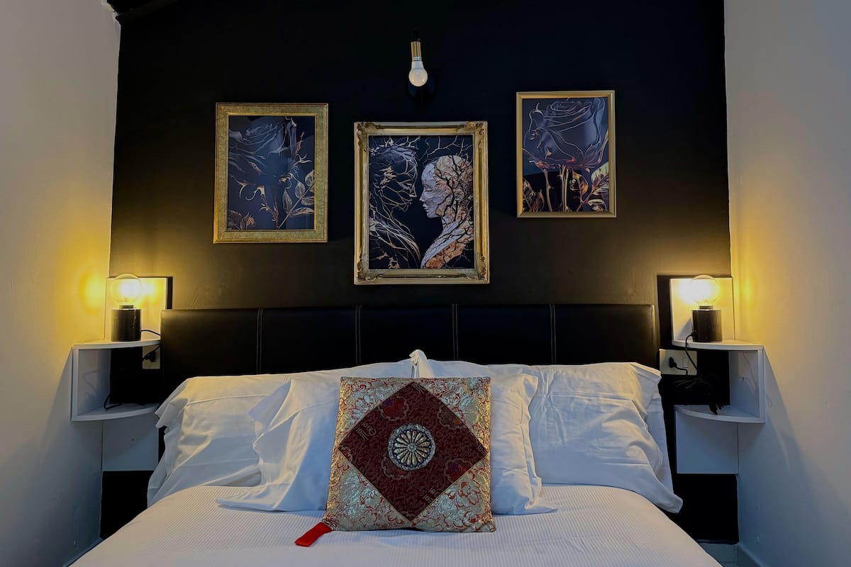 Cama bem arrumada com lençóis brancos e almofada decorativa, ladeada por duas mesas de cabeceira com abajures, em frente a uma parede escura com três obras de arte emolduradas.