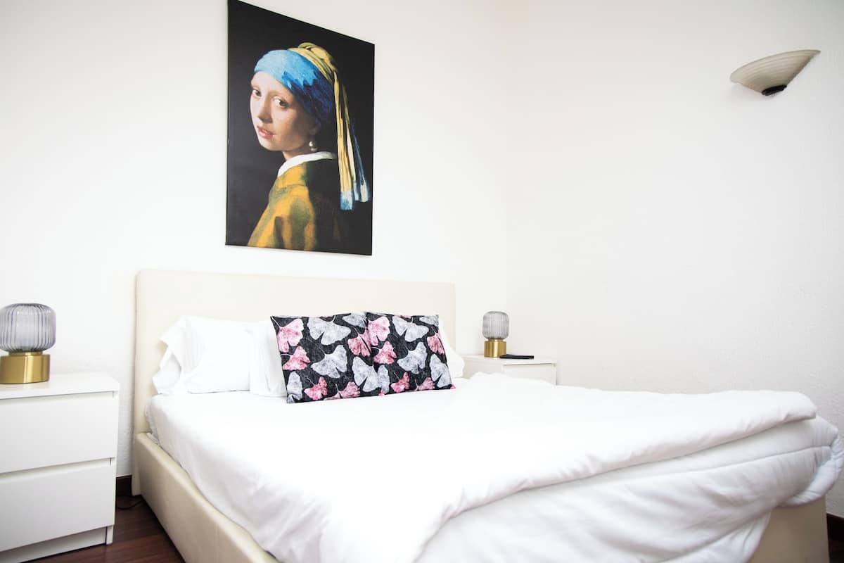 Quarto de hotel com cama, roupa de cama branca, almofada floral, pintura de uma mulher com lenço azul e abajur.