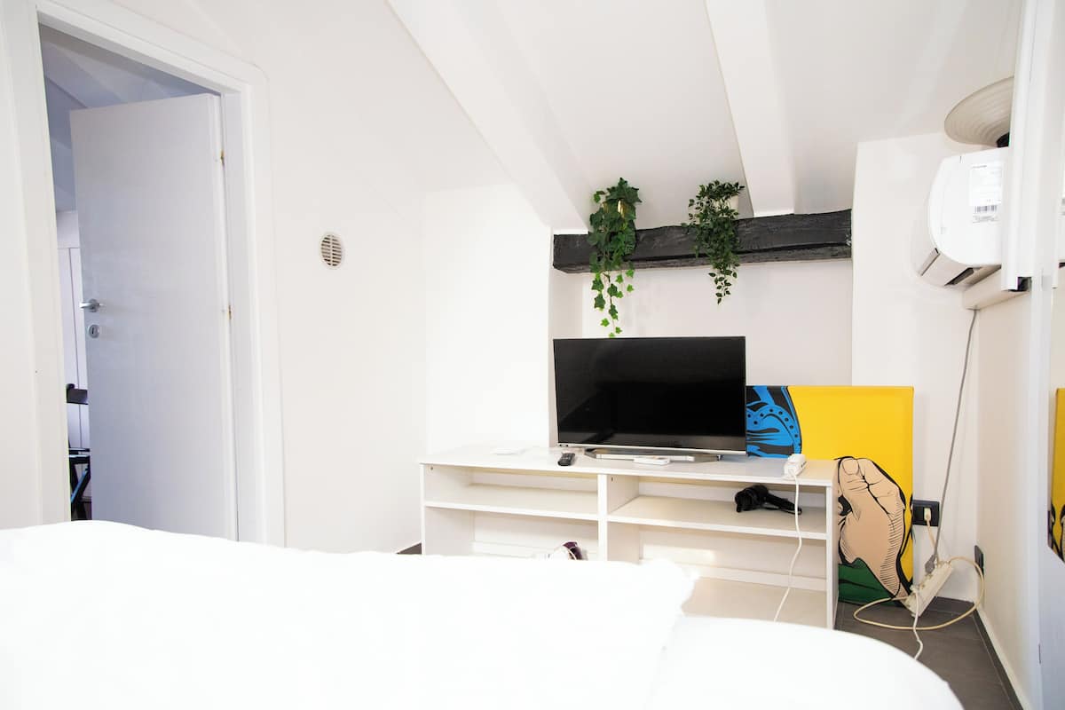 Quarto moderno com cama branca, TV em suporte branco, obra de arte amarela e planta na parede.
