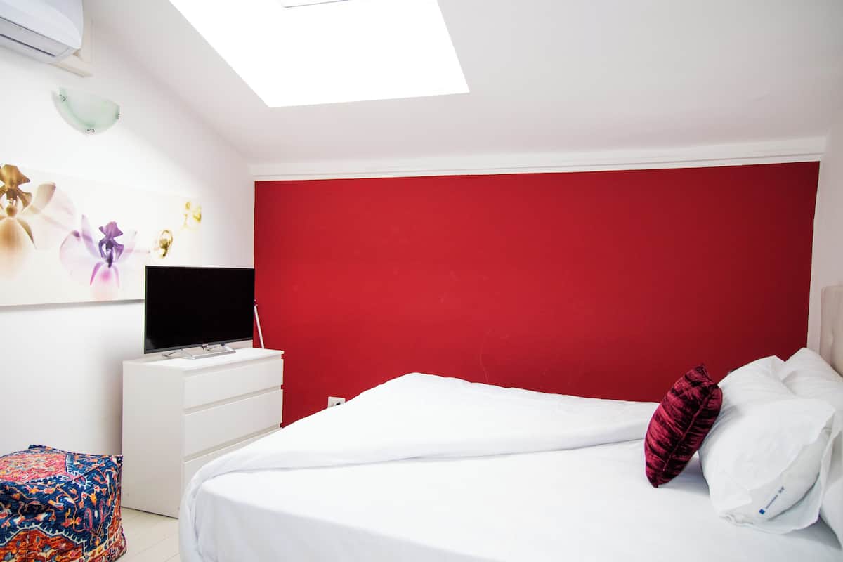 Quarto com parede em destaque vermelha, cama com roupa de cama branca, televisão sobre uma cômoda branca e um claraboia.