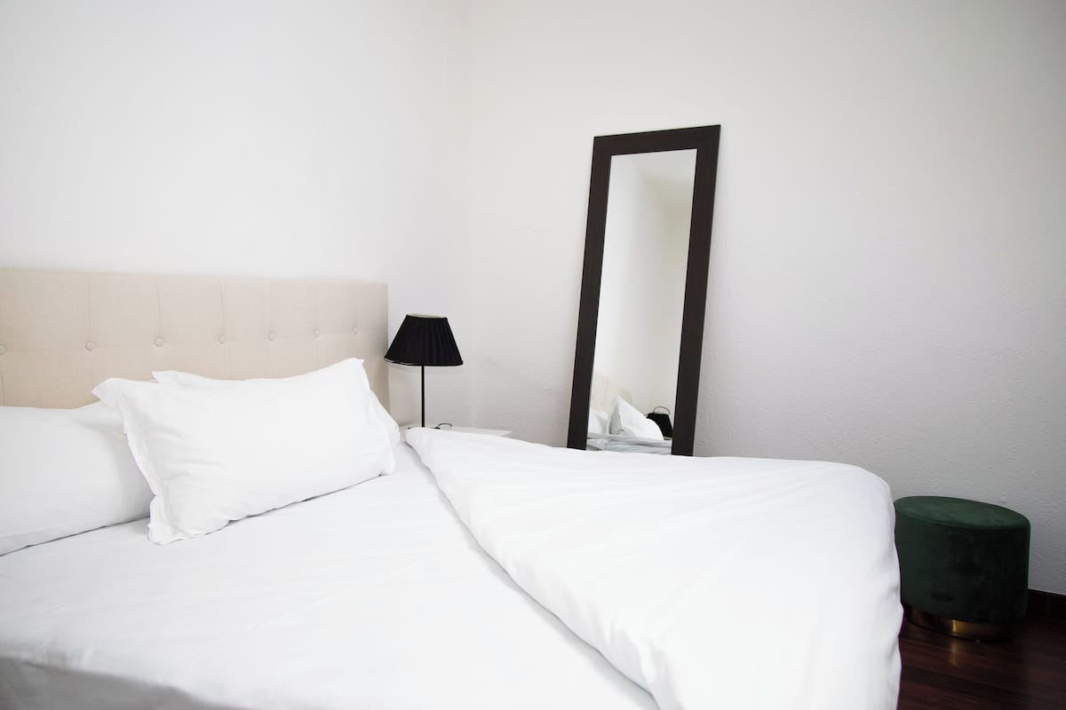 Cama bem arrumada com lençóis e almofadas brancos, uma luminária preta, um espelho grande e um banquinho verde.