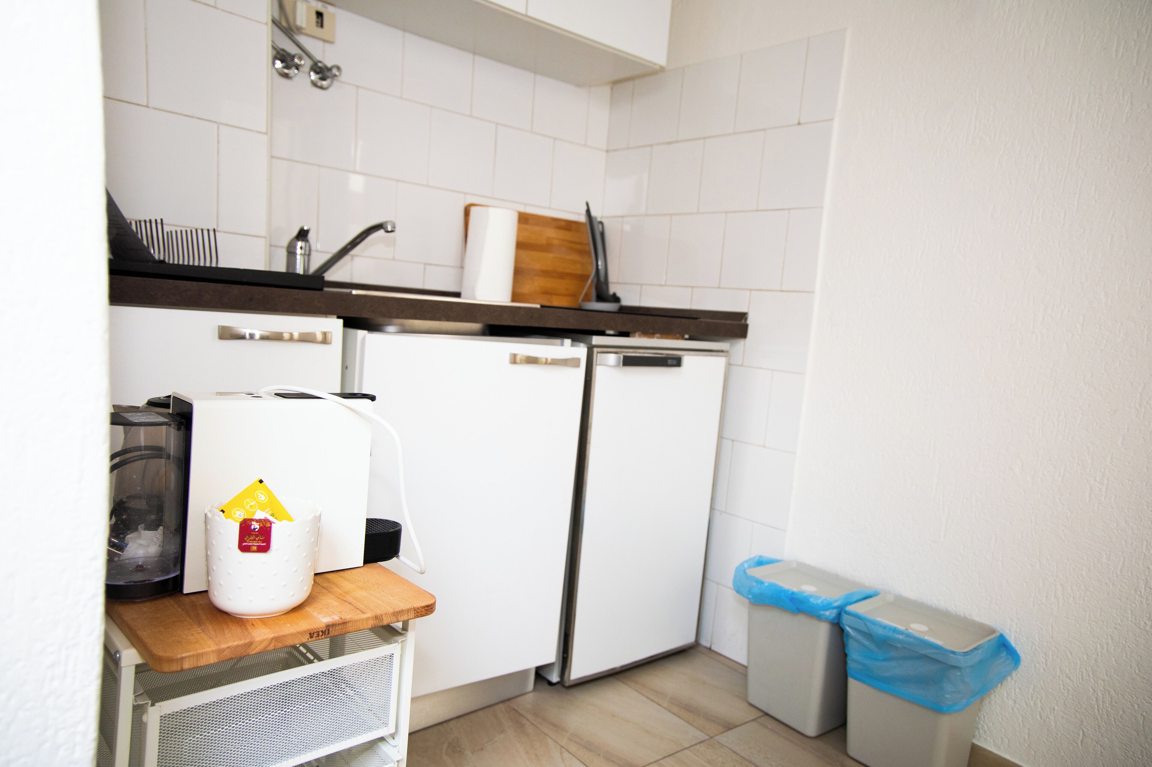 A compact kitchen with a refrigerator, a coffee maker, and a trash bin.
