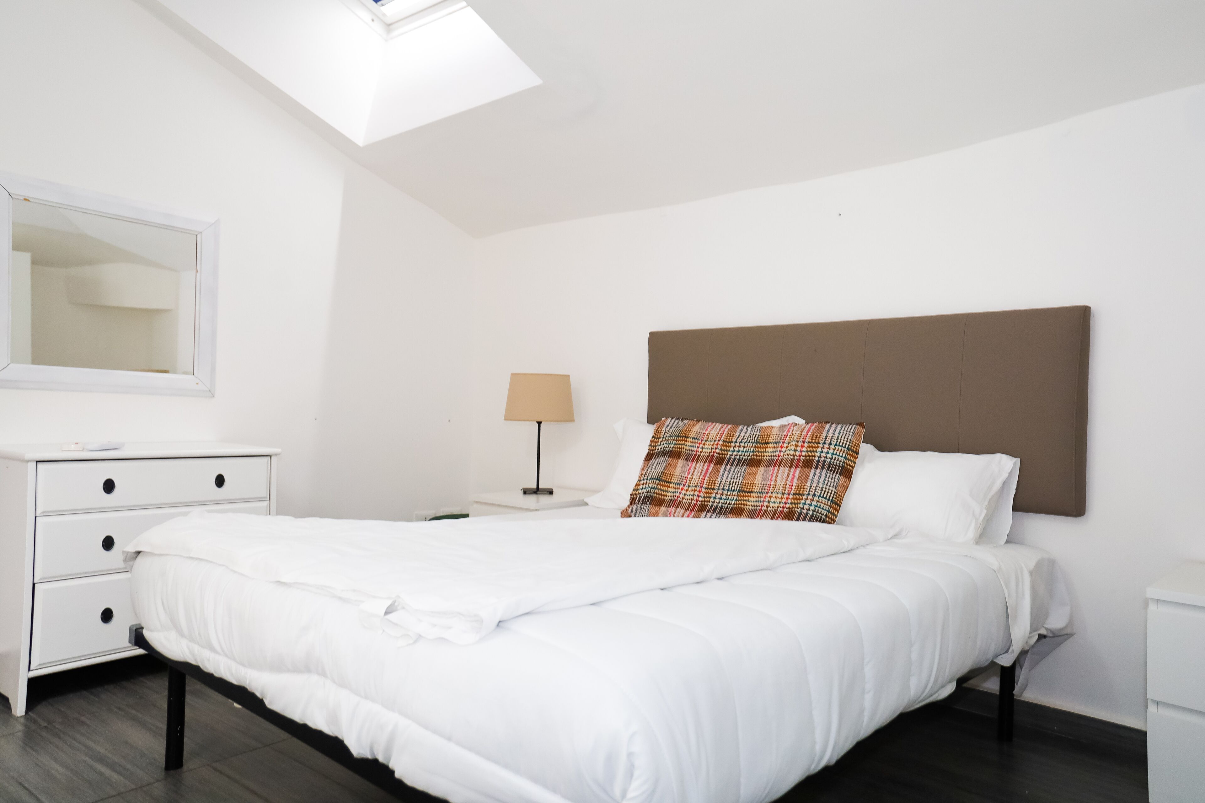 Una cama bien hecha con ropa de cama blanca, cabecera marrón y almohada de cuadros. Hay una mesita de noche blanca con una lámpara y un espejo en la pared.