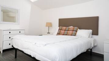 Una cama bien hecha con ropa de cama blanca, cabecera marrón y almohada de cuadros. Hay una mesita de noche blanca con una lámpara y un espejo en la pared.