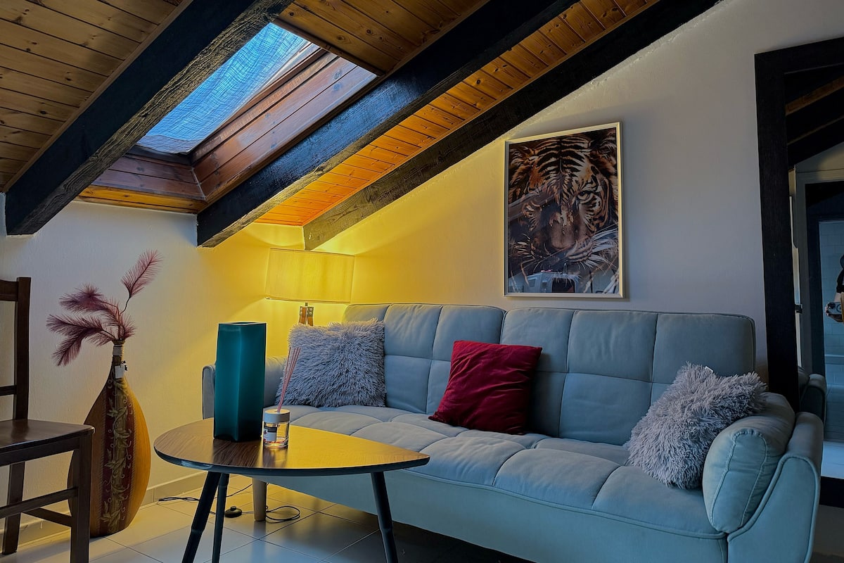 Uma sala de estar moderna com teto alto, vigas de madeira, claraboia, sofá com almofadas, mesa pequena, vaso decorativo e quadro de uma tigresa emoldurada.