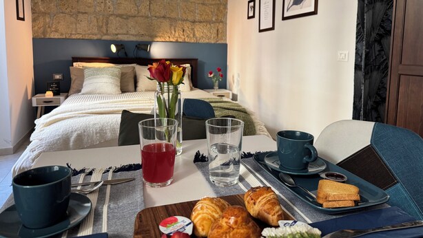 Una mesa de desayuno ordenada con croissants, un vaso de jugo rojo, una taza de café y una bandeja con tostadas y mermelada. Un jarrón con flores está sobre la cama, y la habitación tiene una pared de piedra y obras de arte enmarcadas.