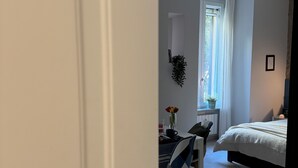 Un dormitorio con cama, escritorio y silla. Hay una ventana con cortinas, una planta y un cuadro en la pared.