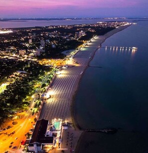 Ein Luftbild einer Küstenstadt in der Abenddämmerung mit einem langen Strand, einem Steg und einer Straße, die zu einem Gebäude mit Pool führt.