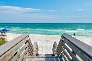 A wooden boardwalk leading to a beach with clear blue water and a few people in the distance.