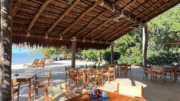 Un área de comedor al aire libre con mesas y sillas de madera, techo de paja y vista a la playa.